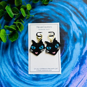 May include: Black cat face earrings with blue eyes and pink noses. The earrings are on a white card with the text "MerakiCreations Handmade Jewelry" and "Let your confidence shine pretty girl #MerakiCreationsbyNeida".