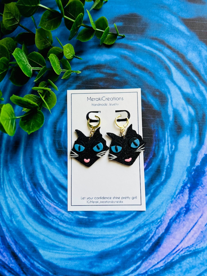 May include: Black cat face earrings with blue eyes and pink noses. The earrings are on a white card with the text "MerakiCreations Handmade Jewelry" and "Let your confidence shine pretty girl #MerakiCreationsbyNeida".