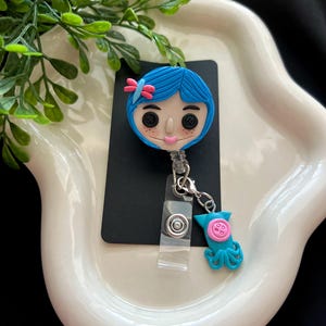 May include: A blue and pink character badge reel with a matching charm. The badge reel features a cartoon character with blue hair, button eyes, and a pink bow. The charm is a teal octopus with a pink button.