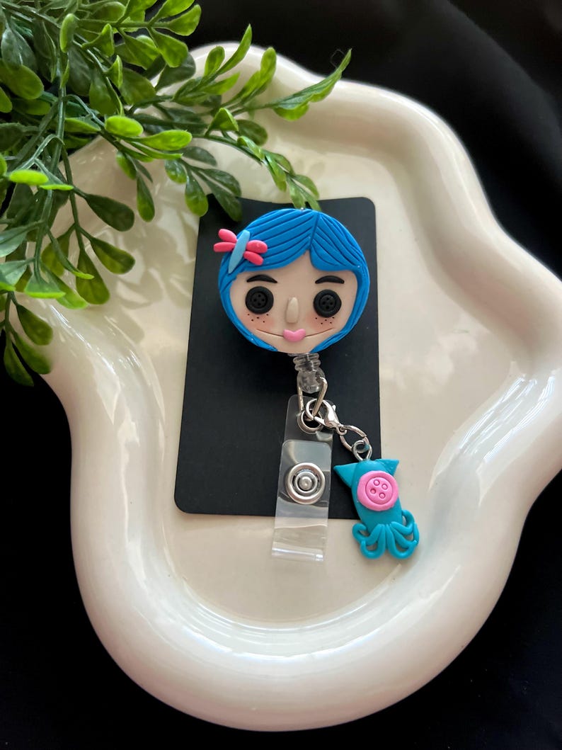 May include: A blue and pink character badge reel with a matching charm. The badge reel features a cartoon character with blue hair, button eyes, and a pink bow. The charm is a teal octopus with a pink button.
