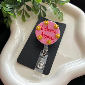May include: A pink badge reel with the words "Welcome home!" in red script, surrounded by yellow flower accents. The reel has a clear plastic strap and a metal clip for attachment.