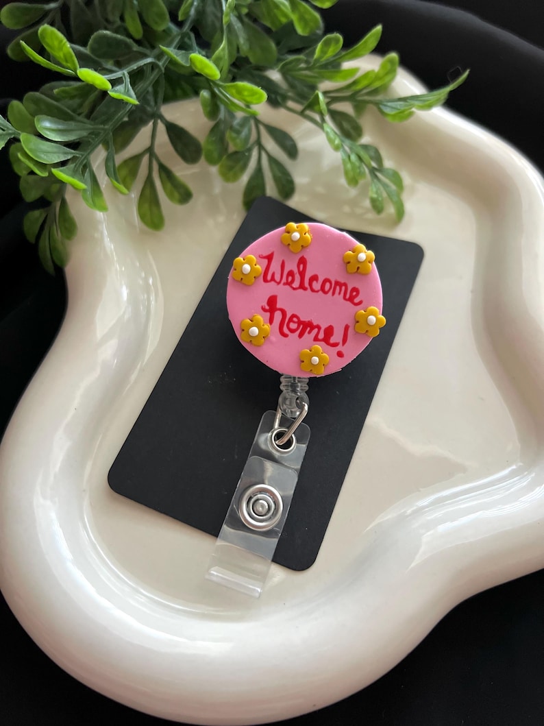 May include: A pink badge reel with the words "Welcome home!" in red script, surrounded by yellow flower accents. The reel has a clear plastic strap and a metal clip for attachment.