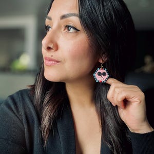 May include: A woman wearing a black top with a pair of pink and blue hexagonal earrings. The earrings have a geometric design with black and white accents.