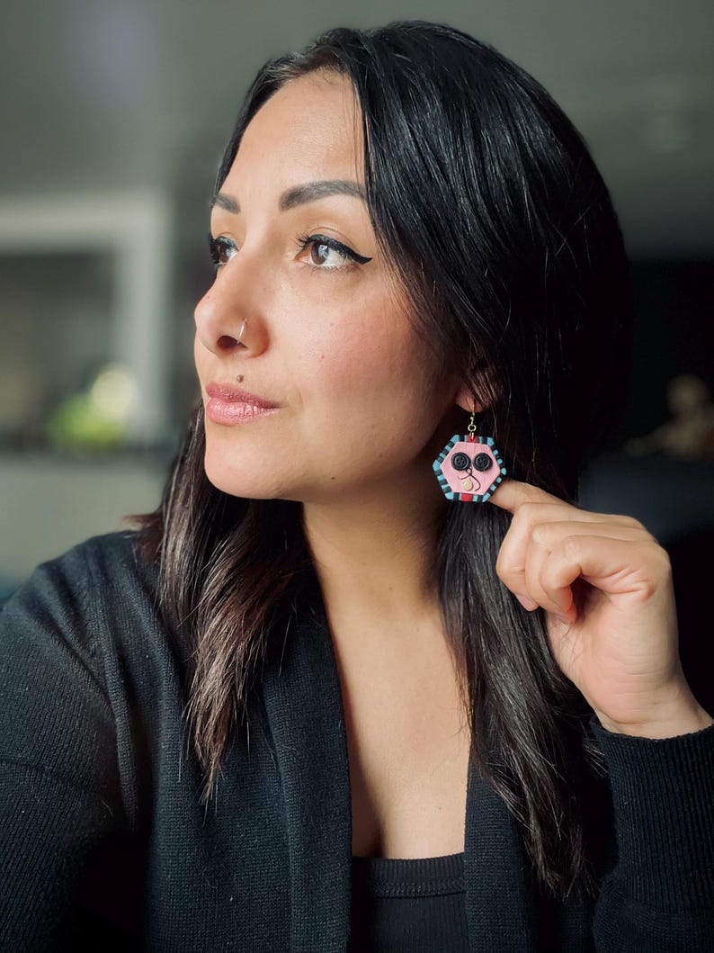 May include: A woman wearing a black top with a pair of pink and blue hexagonal earrings. The earrings have a geometric design with black and white accents.