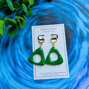 May include: A pair of green triangle-shaped earrings with a gold hoop and a gold hook. The earrings have a circle cut out of the center. The earrings are on a white card with the text "MerakiCreations Handmade Jewelry" and "Let your confidence shine pretty girl! @MerakiCreationsbyneida".