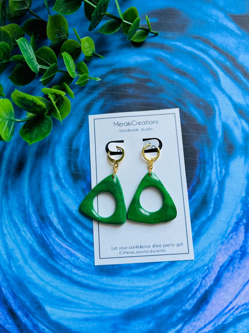 May include: A pair of green triangle-shaped earrings with a gold hoop and a gold hook. The earrings have a circle cut out of the center. The earrings are on a white card with the text "MerakiCreations Handmade Jewelry" and "Let your confidence shine pretty girl! @MerakiCreationsbyneida".