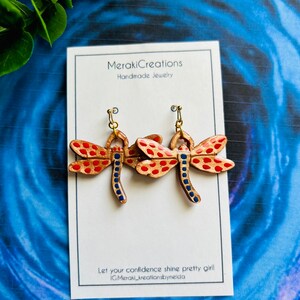 May include: A pair of gold dragonfly earrings with red and blue accents. The earrings are handmade and feature a polka dot pattern.