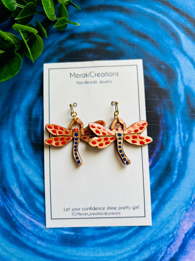 May include: A pair of gold dragonfly earrings with red and blue accents. The earrings are handmade and feature a polka dot pattern.