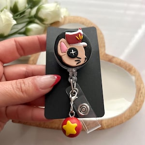 May include: A black badge reel with a cartoon mouse wearing a hat. The mouse has a black button eye. A red and yellow star charm hangs from the reel.