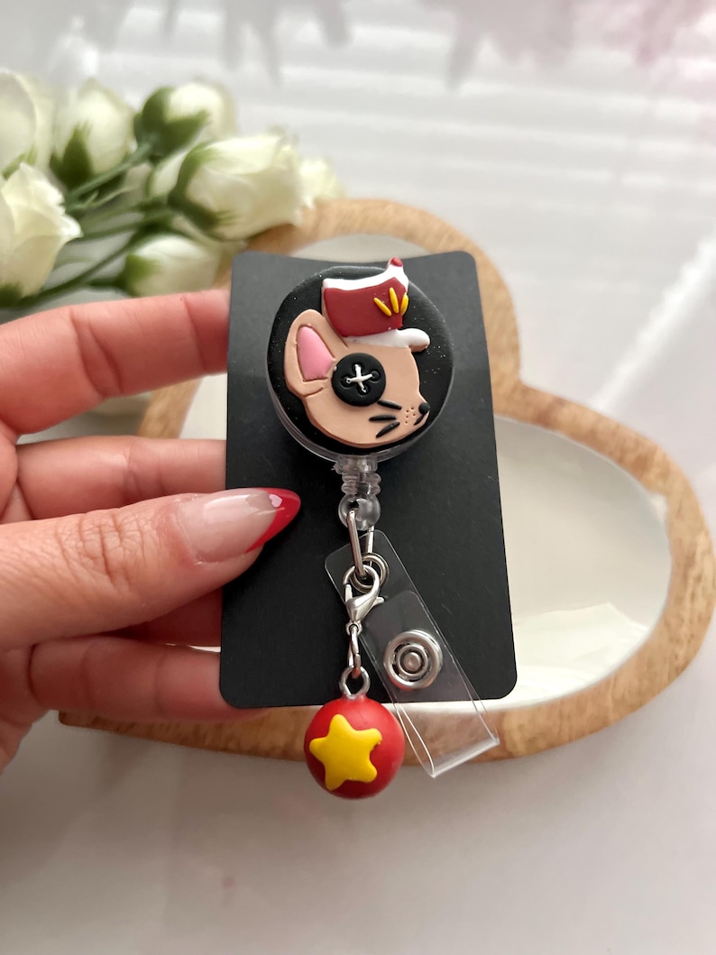 May include: A black badge reel with a cartoon mouse wearing a hat. The mouse has a black button eye. A red and yellow star charm hangs from the reel.