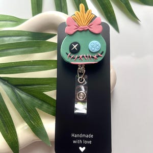 May include: A green and blue character with a pink bow and yellow hair, attached to a black retractable badge holder. The holder has the text "Handmade with love" and a small heart.