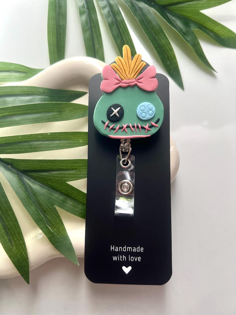 May include: A green and blue character with a pink bow and yellow hair, attached to a black retractable badge holder. The holder has the text "Handmade with love" and a small heart.