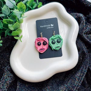 May include: A pair of handmade earrings featuring a pink and green face design with black button eyes and a cross on the forehead. The earrings are on a white plate with a small card that says "Handmade with love".