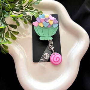 May include: A green badge reel with a heart-shaped flower arrangement on a black background. The arrangement features pastel-colored hearts. A pink and white swirl charm hangs below. The reel is on a white, wavy tray.