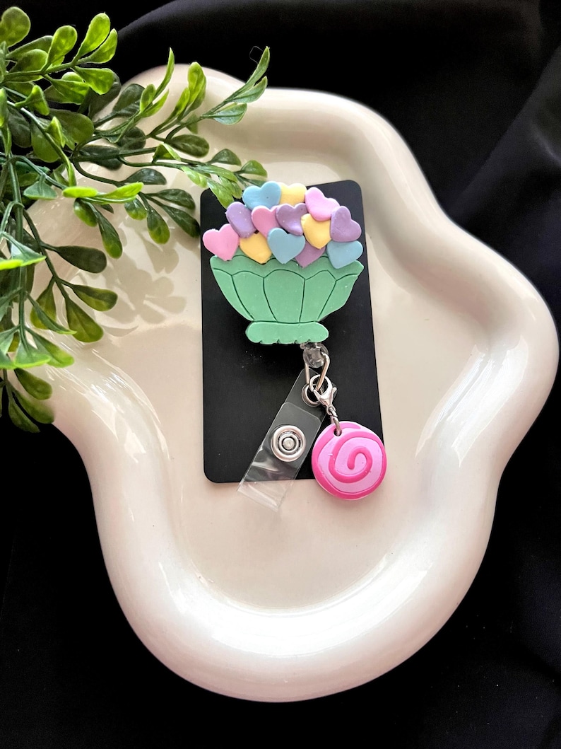 May include: A green badge reel with a heart-shaped flower arrangement on a black background. The arrangement features pastel-colored hearts. A pink and white swirl charm hangs below. The reel is on a white, wavy tray.