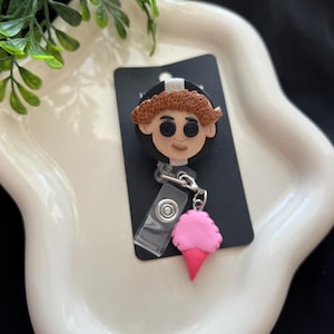 May include: A black badge reel featuring a character with button eyes and brown hair. A pink ice cream cone charm hangs from the reel. The badge reel is on a white, wavy tray with green foliage.
