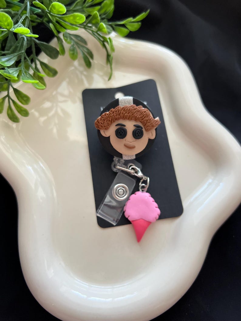 May include: A black badge reel featuring a character with button eyes and brown hair. A pink ice cream cone charm hangs from the reel. The badge reel is on a white, wavy tray with green foliage.