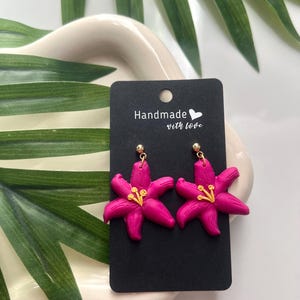 May include: A pair of pink flower earrings with gold accents. The earrings are handmade with love and are displayed on a black card with the text "Handmade with love".