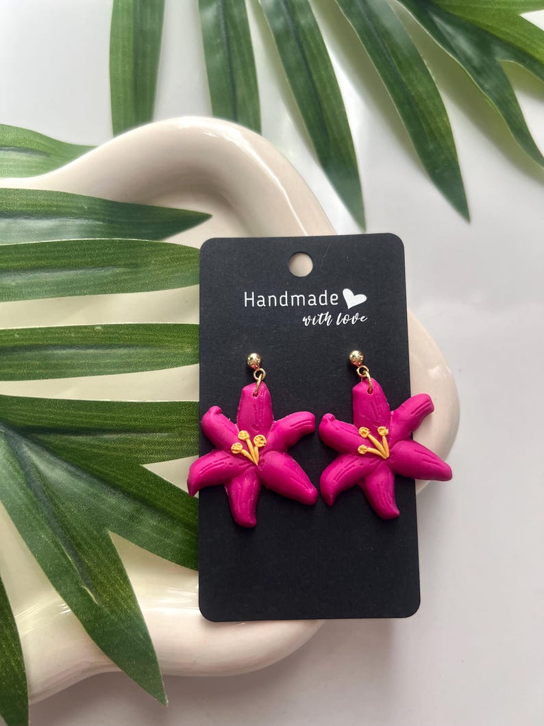May include: A pair of pink flower earrings with gold accents. The earrings are handmade with love and are displayed on a black card with the text "Handmade with love".