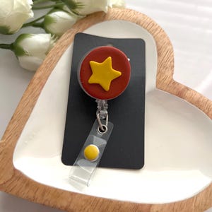 May include: A badge reel with a red circle and a yellow star design. The reel is attached to a black card and has a clear plastic strap with a yellow button. The badge reel is on a white heart-shaped tray.