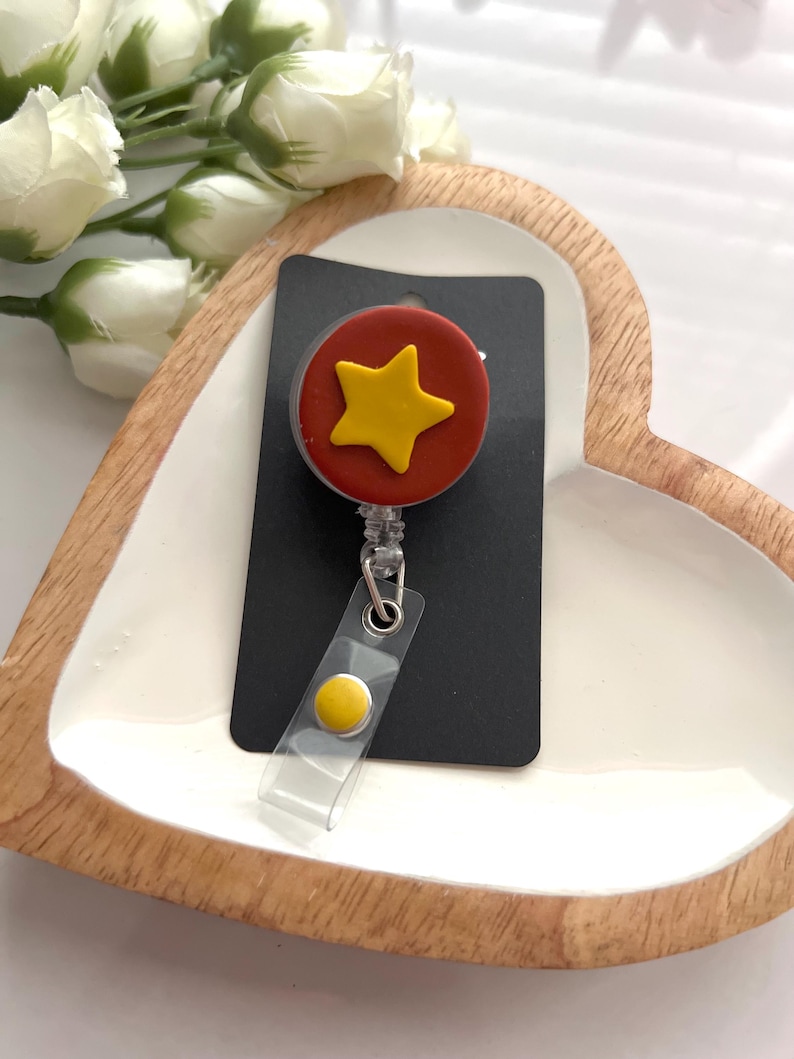May include: A badge reel with a red circle and a yellow star design. The reel is attached to a black card and has a clear plastic strap with a yellow button. The badge reel is on a white heart-shaped tray.