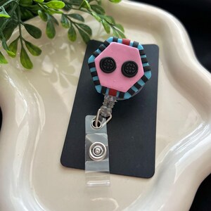 May include: A pink, black, and blue badge reel with a button-like design. The reel has a clear plastic clip and a retractable cord. The design is a hexagon shape with two black button-like features.