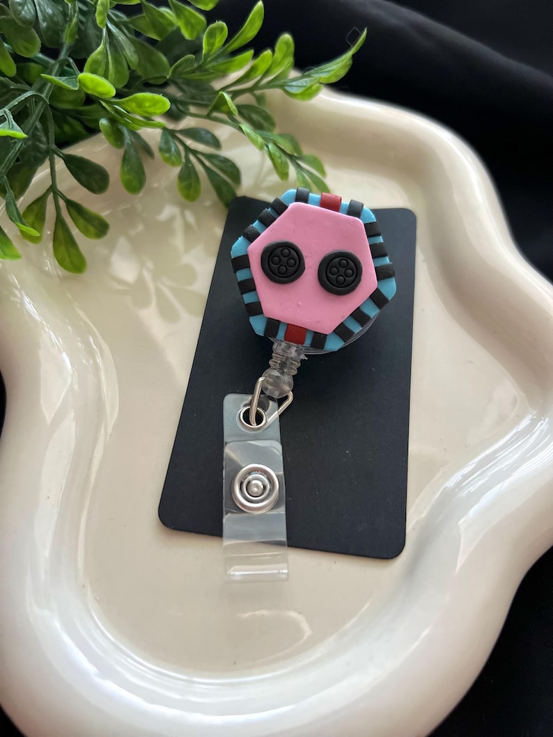 May include: A pink, black, and blue badge reel with a button-like design. The reel has a clear plastic clip and a retractable cord. The design is a hexagon shape with two black button-like features.