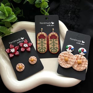 May include: A collection of handmade earrings displayed on black cards. The earrings include red flower studs, stained glass-style drop earrings, and waffle-shaped earrings. Each card is labeled "Handmade with love."
