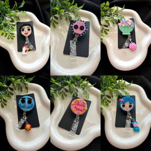 May include: A collection of six handmade badge reels with various character designs. Each reel features a unique character, including a girl, a rooster, and a character with the text "Welcome home!". The reels are attached to black rectangular cards.