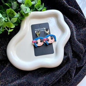 Pu&ograve; includere: Orecchini fatti a mano con un design a volto di cartone animato, capelli blu, fiocchi rosa e occhi a bottone. Gli orecchini sono su una carta nera con cerchi dorati. La carta &egrave; su un piatto bianco.