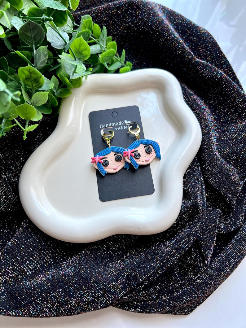 Pu&ograve; includere: Orecchini fatti a mano con un design a volto di cartone animato, capelli blu, fiocchi rosa e occhi a bottone. Gli orecchini sono su una carta nera con cerchi dorati. La carta &egrave; su un piatto bianco.