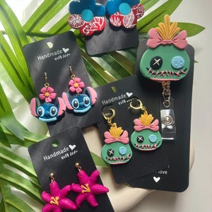 May include: Four pairs of handmade polymer clay earrings featuring a variety of designs, including a blue flower with white leaves, a blue character with a pink flower, a green character with a pink bow, and a pink flower. The earrings are displayed on black cards with the text "Handmade with love".