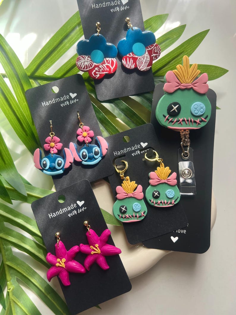 May include: Four pairs of handmade polymer clay earrings featuring a variety of designs, including a blue flower with white leaves, a blue character with a pink flower, a green character with a pink bow, and a pink flower. The earrings are displayed on black cards with the text "Handmade with love".
