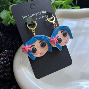 Pu&ograve; includere: Orecchini fatti a mano raffiguranti un personaggio dei cartoni animati con capelli blu, occhi a bottone e un fiocco rosa. Gli orecchini sono appesi a cerchi dorati e a una carta nera con la scritta "Handmade with love."