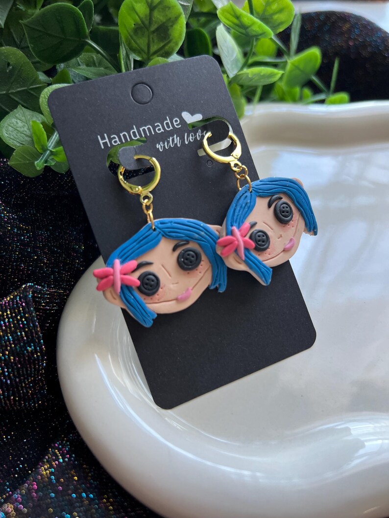 Pu&ograve; includere: Orecchini fatti a mano raffiguranti un personaggio dei cartoni animati con capelli blu, occhi a bottone e un fiocco rosa. Gli orecchini sono appesi a cerchi dorati e a una carta nera con la scritta "Handmade with love."