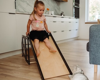 May include: A young girl slides down a wooden slide attached to a black wooden climbing frame. A gray cat watches from the floor.