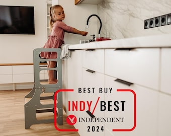 May include: A child stands on a gray wooden step stool in a kitchen. The stool has three steps and a safety rail. The child is reaching up to a kitchen sink. The text "BEST BUY INDY/BEST INDEPENDENT 2024" is visible in the lower right corner of the image.