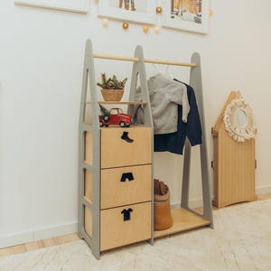Puede incluir: Un perchero de madera clara y gris con tres cajones apilados con tiradores negros, una barra para colgar ropa y un estante abierto. La unidad está decorada con una pequeña cesta tejida con vegetación y un camión de juguete rojo en el estante superior.