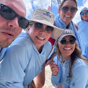 May include: Four people are smiling for a photo. Three of them are wearing light blue shirts with a logo. One person is wearing a tan hat with the text "Bitter End Yacht Club".