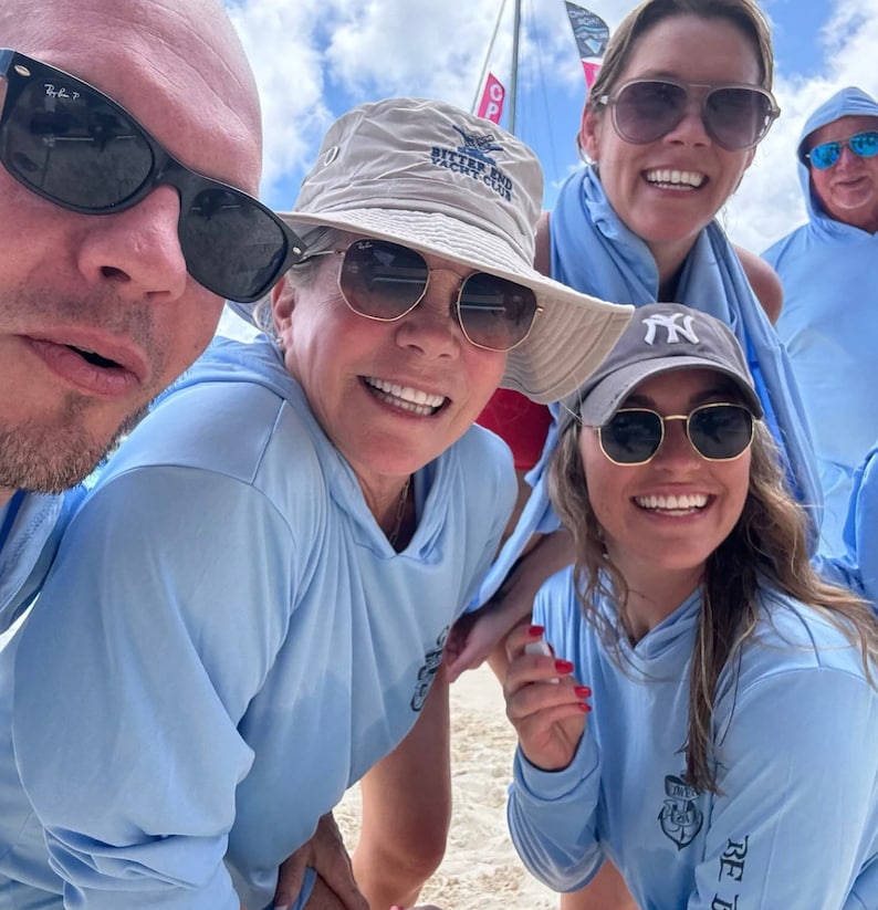May include: Four people are smiling for a photo. Three of them are wearing light blue shirts with a logo. One person is wearing a tan hat with the text "Bitter End Yacht Club".