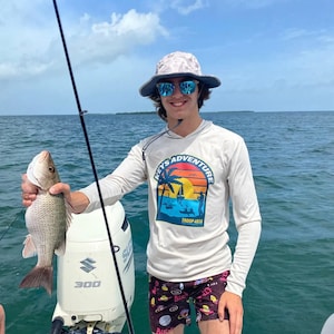 May include: A person wearing a white long-sleeved shirt with a graphic of a palm tree and the text "Keys Adventure Troop 4018" is holding a fish. The person is wearing a grey and white bucket hat and sunglasses. The person is standing on a boat with a white outboard motor.