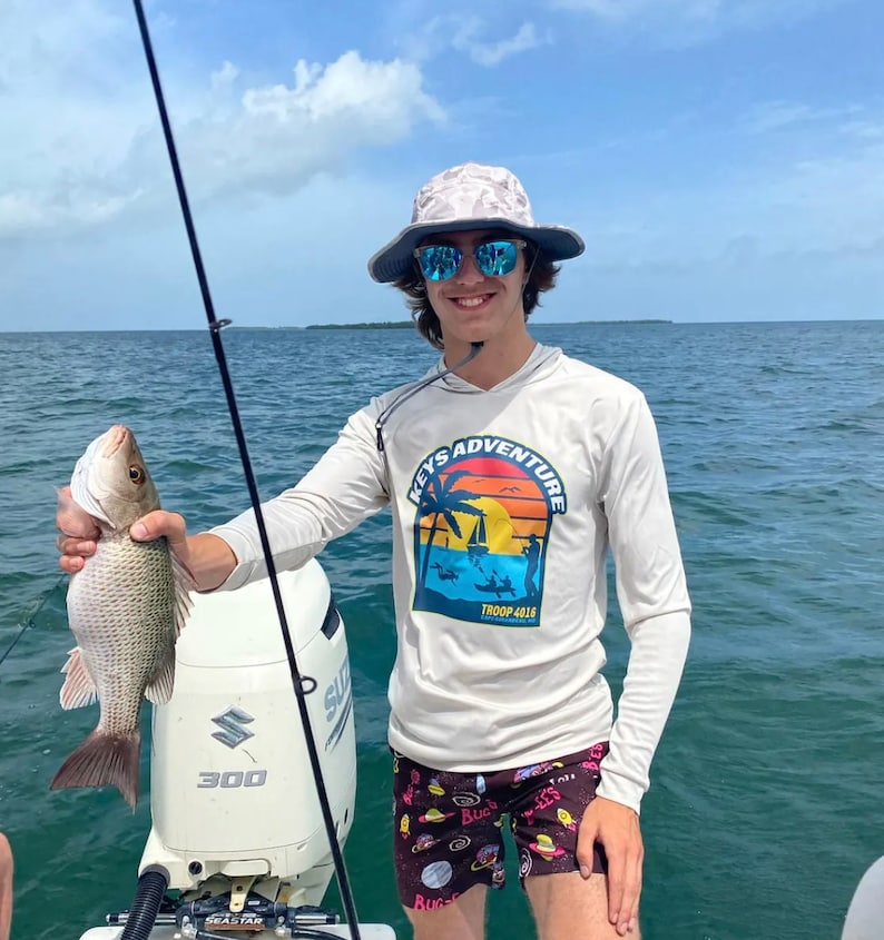 May include: A person wearing a white long-sleeved shirt with a graphic of a palm tree and the text "Keys Adventure Troop 4018" is holding a fish. The person is wearing a grey and white bucket hat and sunglasses. The person is standing on a boat with a white outboard motor.