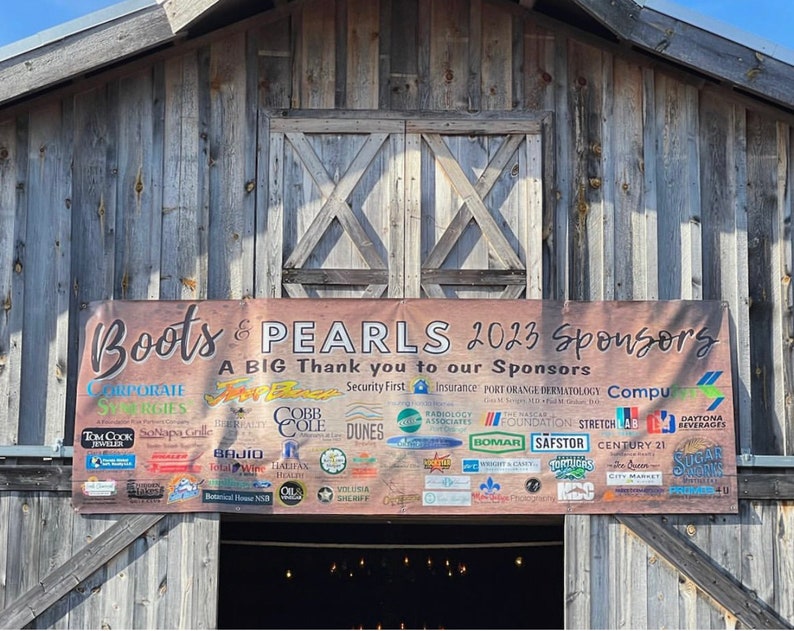 May include: A brown wooden barn with a banner that reads "Boots & PEARLS 2023 Sponsors" and lists a variety of sponsors. The banner is brown with white text and logos.