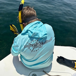 May include: A light blue hooded shirt with a compass design and the text "Latitude Adjustment" printed on the back.