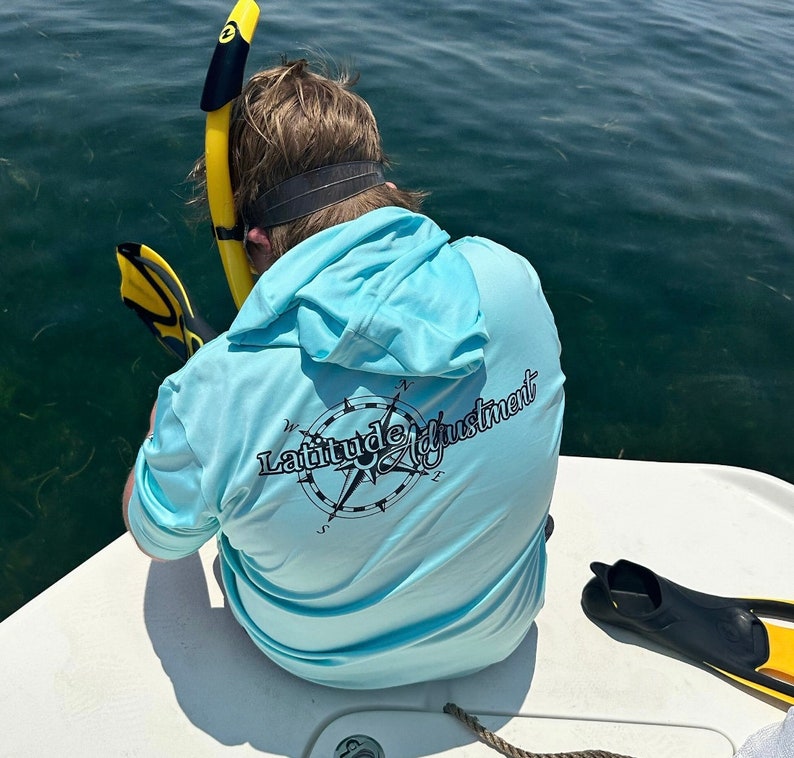 May include: A light blue hooded shirt with a compass design and the text "Latitude Adjustment" printed on the back.