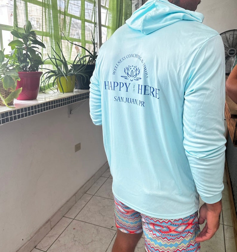 May include: A light blue long-sleeved shirt with a hood. The back of the shirt has a logo that says "Wellness Coaching & Tours" with a lotus flower design and "Happy Here" with the text "San Juan, PR" underneath.