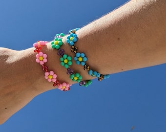 Puede incluir: Tres pulseras de cuentas con dijes en forma de flor. Las pulseras son de color rosa, verde y azul con centros amarillos. Las pulseras están apiladas en la muñeca de una persona.