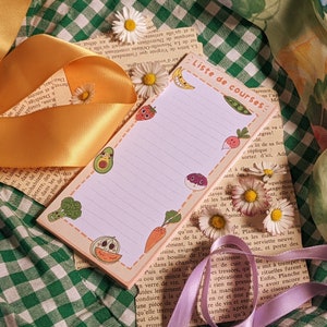 Peut inclure: Un bloc-notes de liste de courses rose et blanc avec le titre manuscrit "Liste de courses" et une liste d'articles en français. Le bloc-notes est décoré de dessins animés colorés de légumes et de fruits.