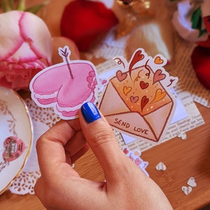 Peut inclure: Deux autocollants sur le thème de la Saint-Valentin. Un autocollant est un biscuit en forme de cœur rose avec une flèche qui le traverse. L'autre autocollant est une enveloppe avec des cœurs et des paillettes qui en sortent. Le texte sur l'enveloppe dit "Envoyer de l'amour".