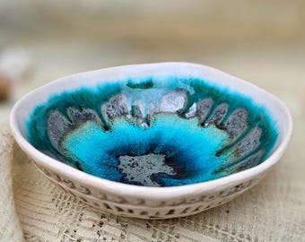 May include: A small, handmade ceramic bowl with a unique design. The interior features a vibrant blend of turquoise, teal, and dark blue hues. The bowl's rim is white, and the exterior has a decorative pattern. A beautiful piece for serving or display.